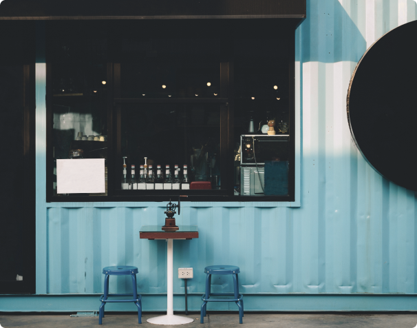 Exterior view of a modern coffee shop featuring a bright blue shipping container design, large windows displaying a variety of beverages, and a small table with two blue chairs in front.
