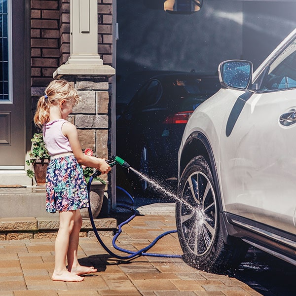 A young girl using a hose to spray the rim of an SUV parked on her driveway.