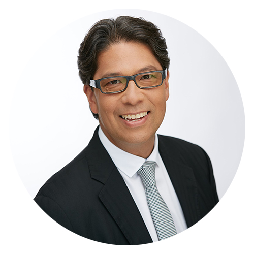 Professional headshot of a smiling man with dark hair and glasses, wearing a black suit and white shirt with a tie, set against a plain white background.