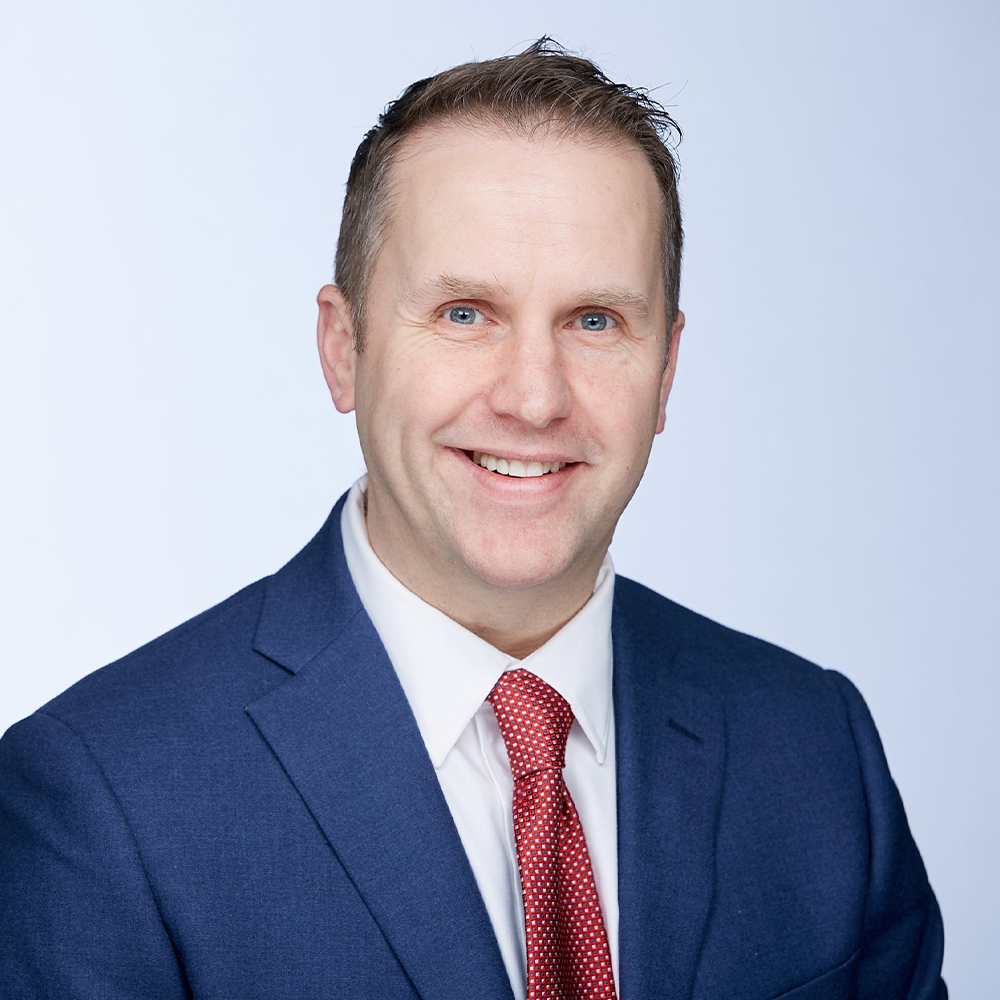 Professional headshot of a man smiling, wearing a navy blue suit with a white shirt and red tie, against a light background. Suitable for corporate or business profiles.