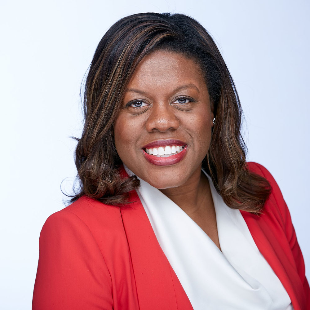 Professional headshot of a smiling woman with dark hair styled in loose waves, wearing a bright red blazer over a white blouse, set against a light background.