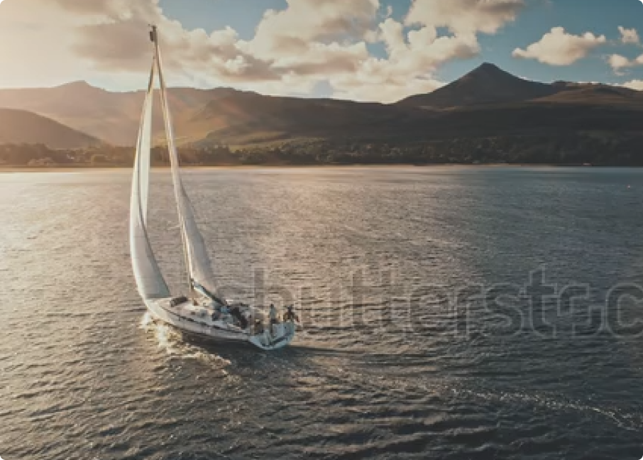 Sailboat with white sails gliding across a serene lake at sunset, surrounded by mountains and a clear sky, creating a picturesque scene for sailing enthusiasts and nature lovers.