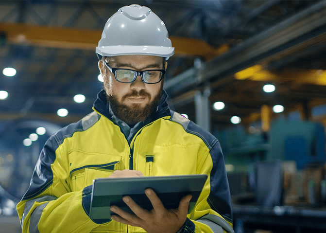 A worker wearing a helmet looks at a tablet in a manufacturing plant.