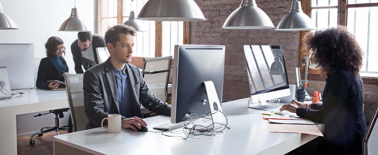 Two office workers sitting at their desktop computers in a modern office with a brick interior.