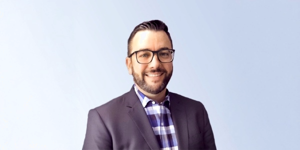 Professional man smiling in a suit and glasses, standing against a light blue background, ideal for business or corporate use.