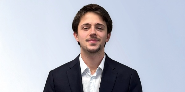 Young man in a formal suit with a white shirt, smiling against a light blue background, representing professionalism and confidence.
