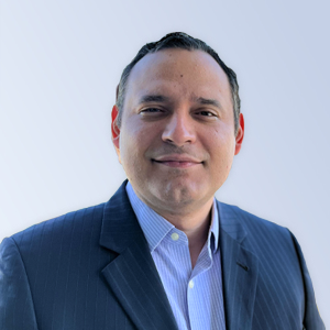 Professional headshot of a man wearing a dark suit and light blue shirt, smiling against a soft gradient background. Ideal for business profiles or professional networking.